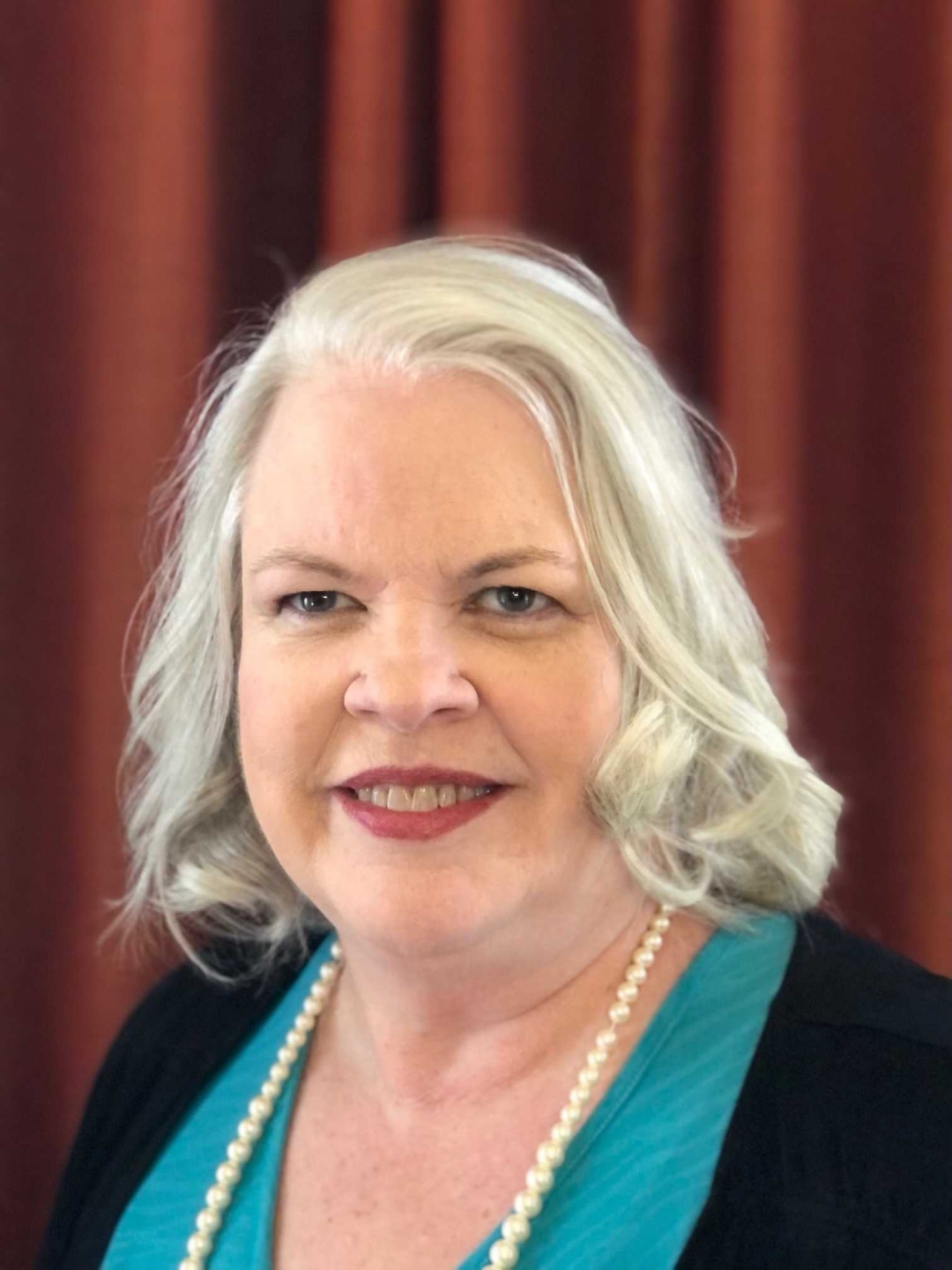 Women with white hair and a blue and black shirt with a pearl necklace and red backdrop. 