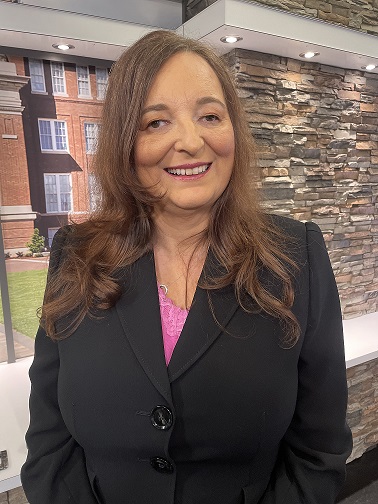 Sylvia Stinson-Perez Standing brown long hair in a black business suit and a pink shirt with stone bricks in the background.