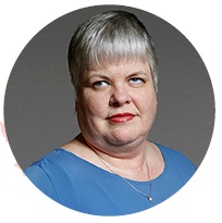 A face picture of Neva a woman with grey hair, a blue shirt, and a gold necklace.