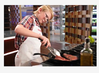 Alfred stands at a stove while cooking 2 steaks in a pan. He is lifting the edge of one with tongs to check its doneness.