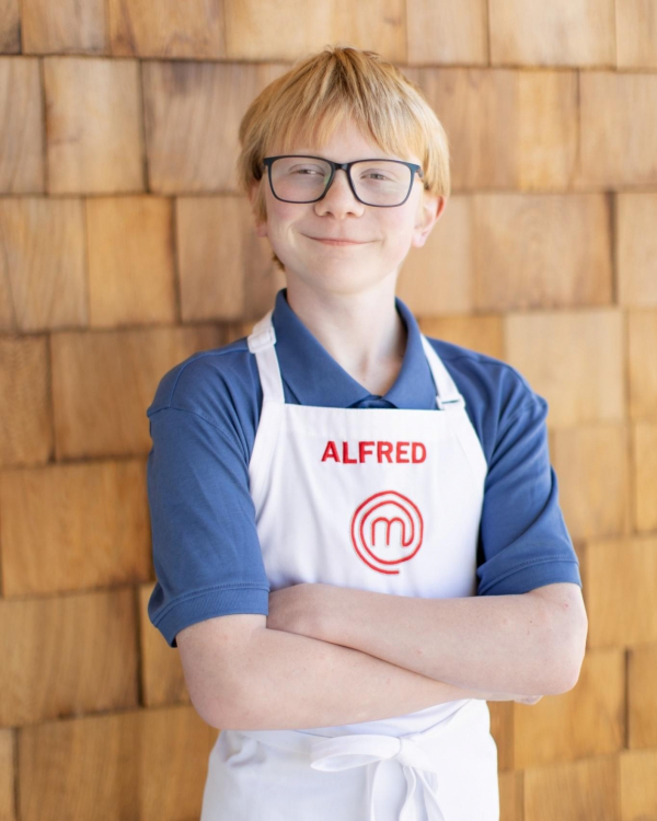 Wearing an apron that says "Alfred" at the top and the masterchef logo below, Alfred smiles at the camera. He has shaggy red hair and wears black-rimmed glasses.