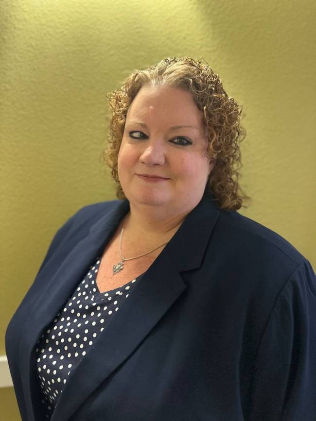 Wearing a navy suit jacket and navy blouse with white polka-dots, Amy smiles at the camera. She has curly reddish-brown hair.