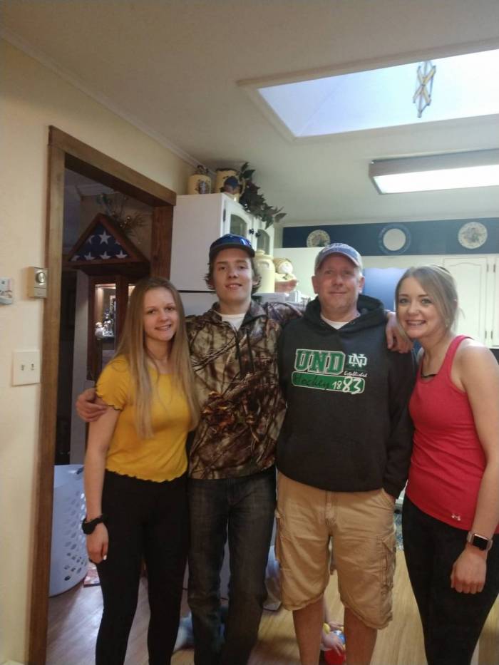 Arik stands in a kitchen with his 3 young adult children - 2 girls and one boy. They stand in a row with their arms around each other.