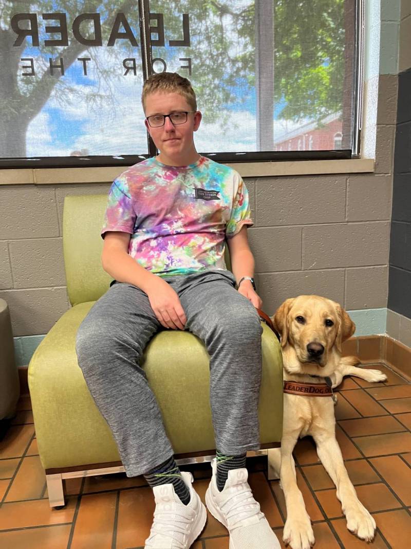 Ethan sits on a chair with a yellow lab seated next to him on the floor.