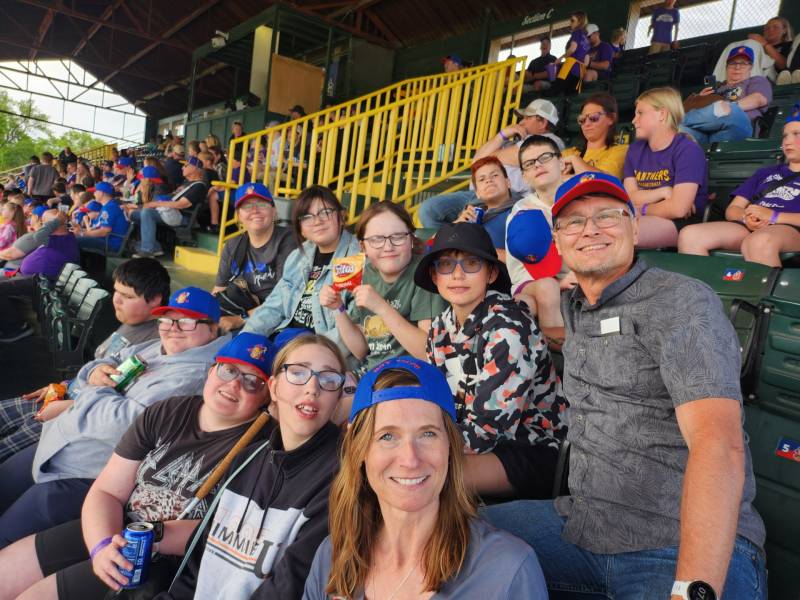 students and staff sit in the stands at a baseball game. they all are looking towards the camera and smiling.