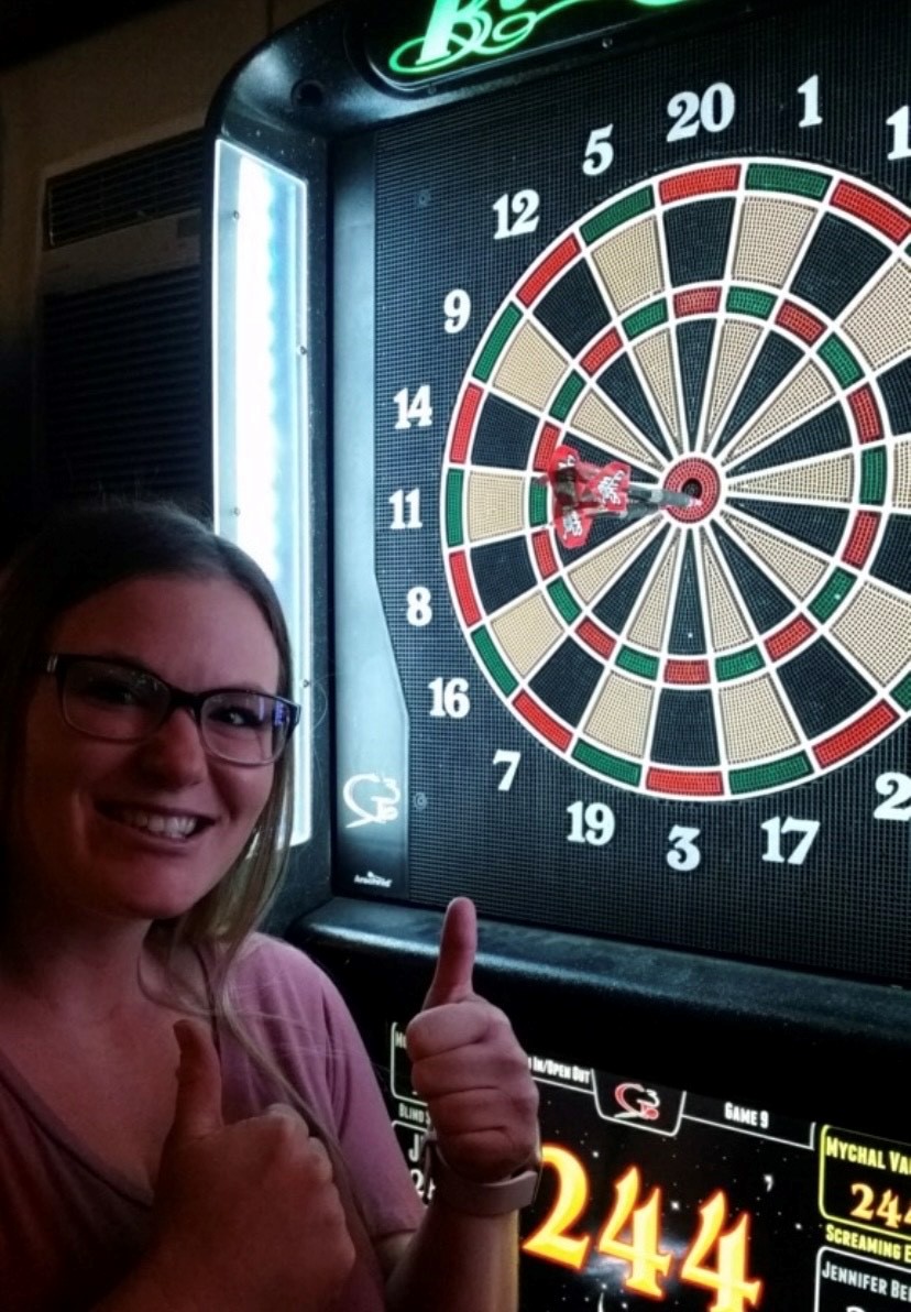 Morgan gives a thumbs up as the dart board behind her displays 3 darts in the bullseye.
