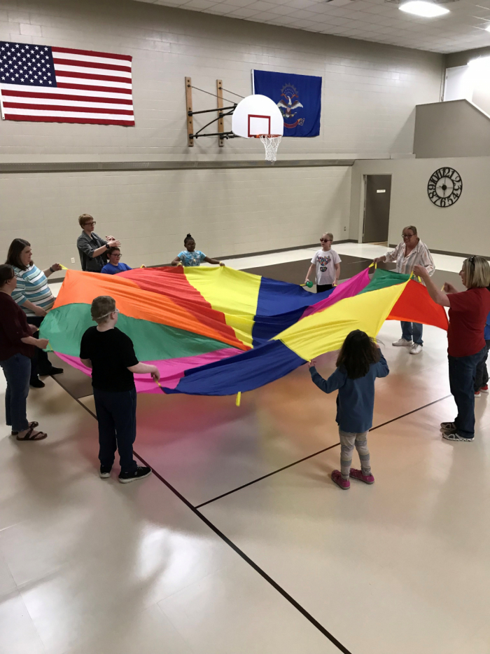 Elementary students and staff members hold onto a large rainbow parachute in the gym at NDVS/SB.