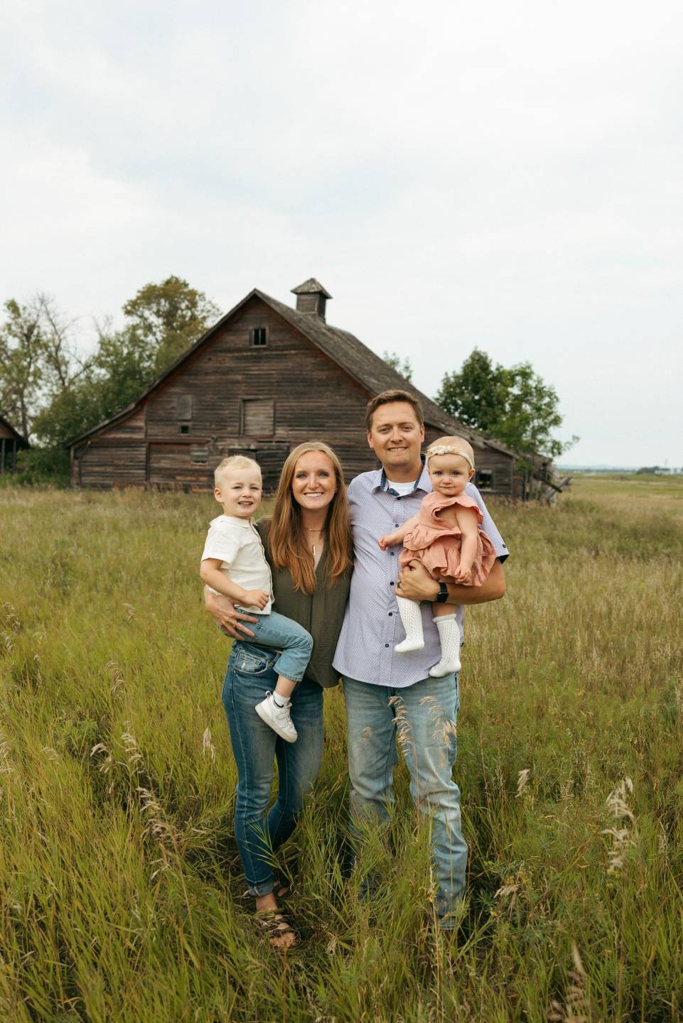 Breanne and her husband stand in a field of grass in front of an old farm building. Breanne holds a boy toddler and her husband holds a baby girl.