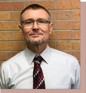 Image of Paul Olson a middle aged man with short hear, glasses, goatee, with a white shirt and red tie standing by a brick wall.