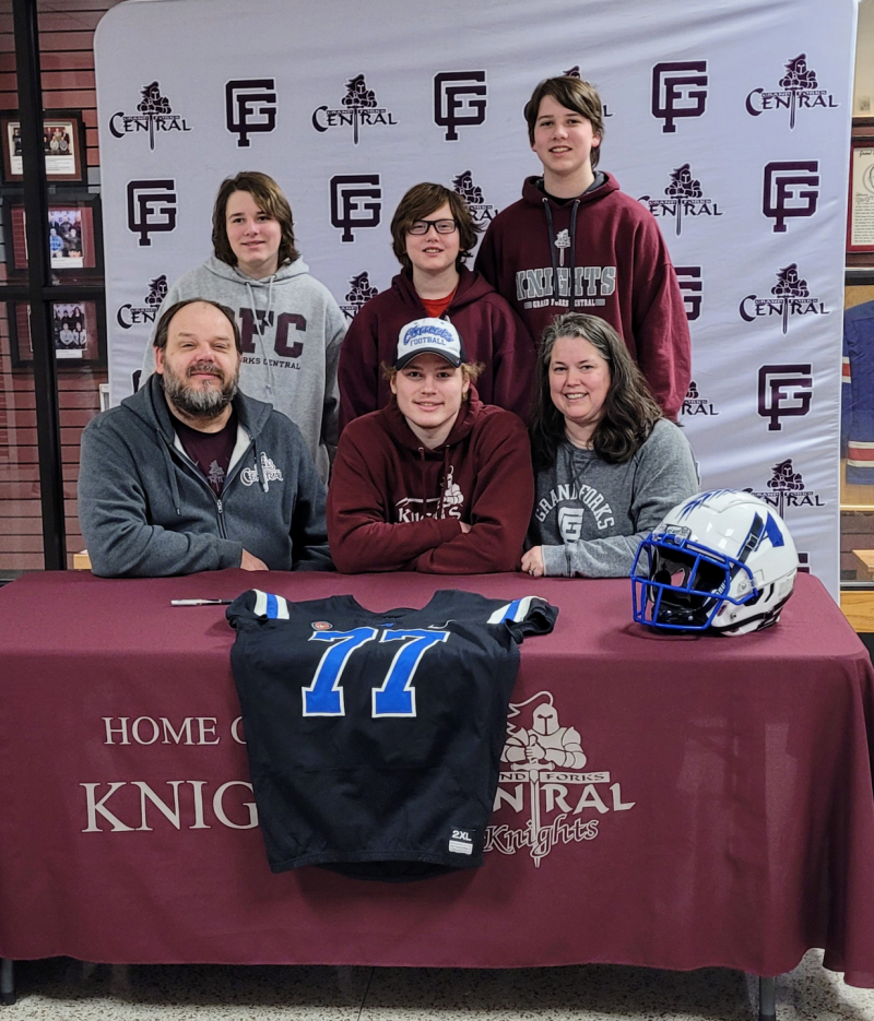 Ryan with his wife and four sons. Ryan, Katie, and Michael are seated behind a table. The other 3 sons are standing behind them. Michael has a Mayville State football jersey on the table in front of him. He has just signed a contract to play football there after high school.